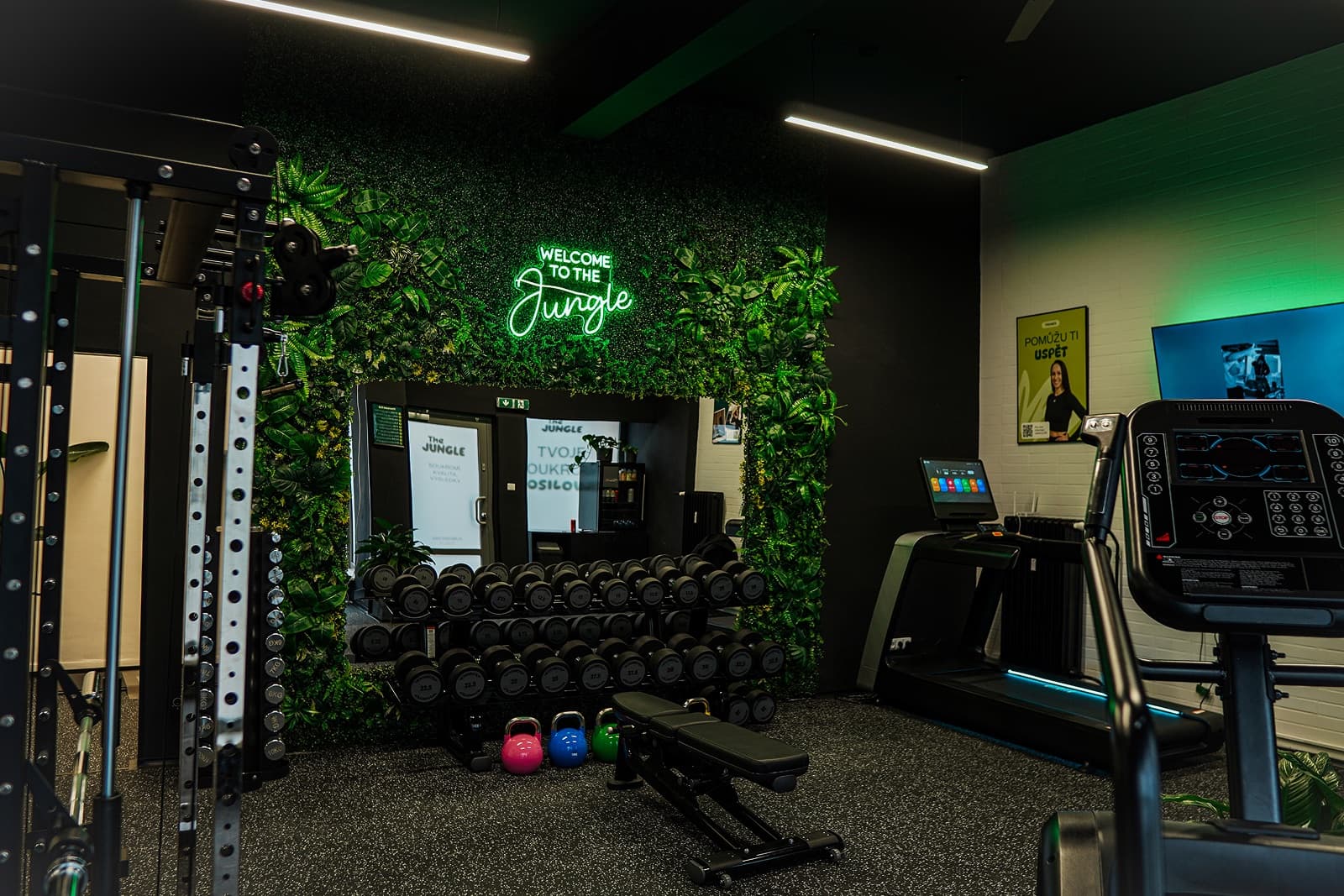 Prázdný interiér posilovny The Jungle se zelenou stěnou z rostlin a neonovým nápisem Welcome to the Jungle, stojanem s jednoručkami, multipress klecí a běžeckým pásem